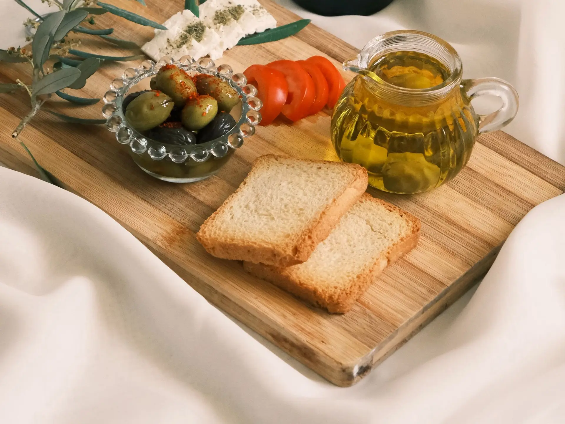 Pan tostado, aceitunas, tomate, queso fresco y una jarra de aceite sobre una tabla