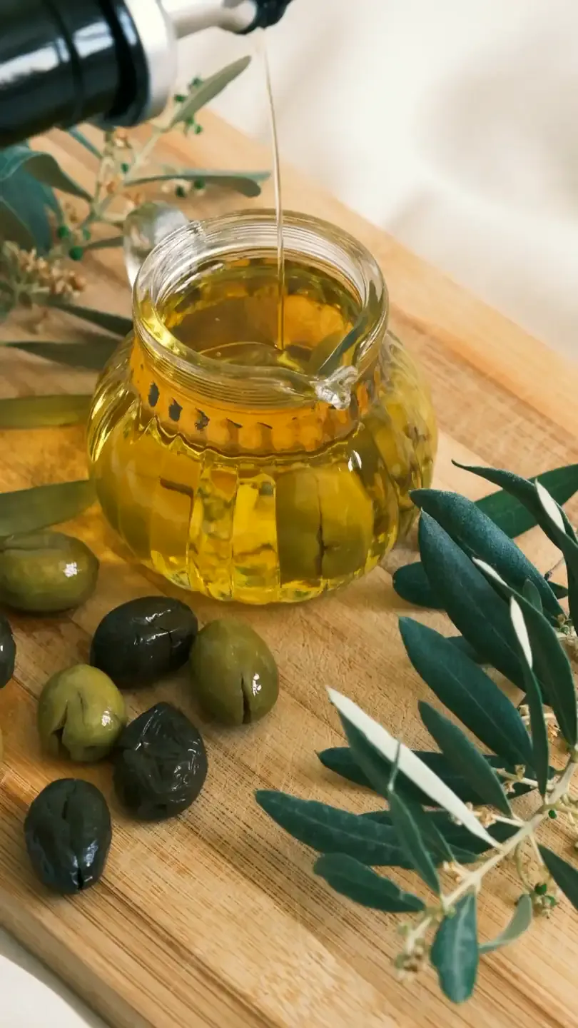 Aceite de oliva cayendo en una jarra de cristal junto a aceitunas y ramas de olivo